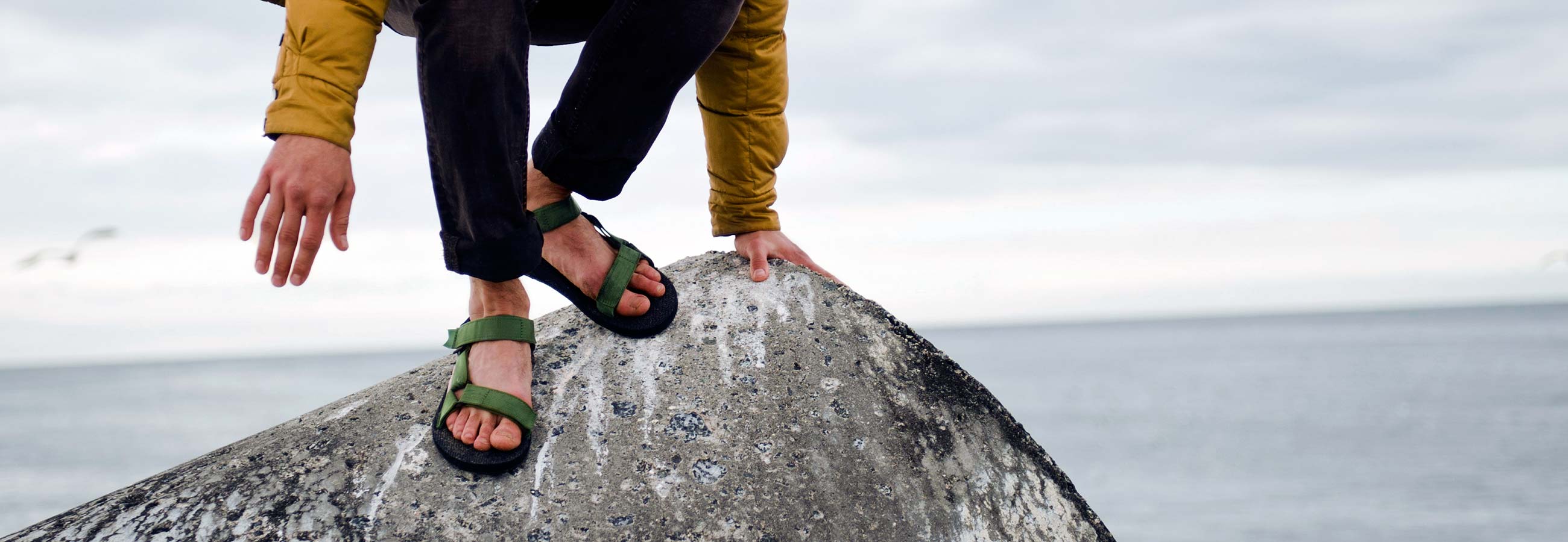 Lower half of person wearing Teva sandals climbing down a boulder overlooking the ocean.